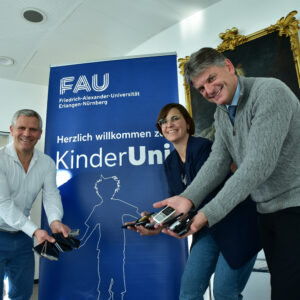 Philipp Weber gemeinsam mit Prof. Hornegger und Laura Kneppe zeigen ausgediente Handys, die für die Kreativ-Spenden gesammelt wurden.